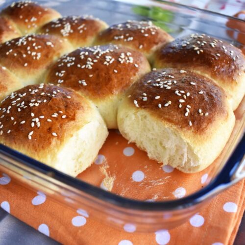 Golden homemade dinner rolls served in a Pyrex baking dish, topped with sesame seeds, with a soft, fluffy texture and shiny, buttery crust