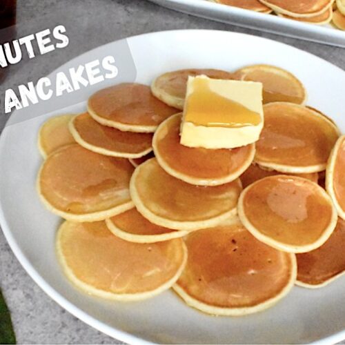 Mini pancakes served on a round white plate, topped with a pat of butter and drizzled with maple syrup.