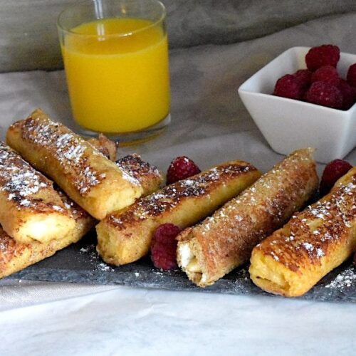 Golden French Toast Roll-Ups, served on a black plate, topped with powdered sugar and fresh raspberries, with a cup of orange juice and a small white bowl of raspberries on the side.