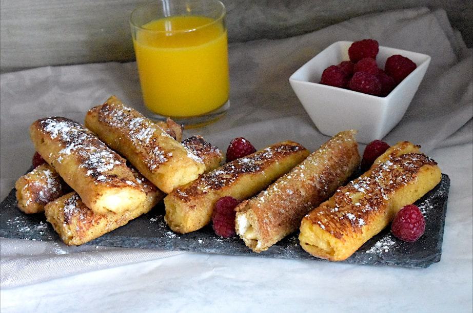 Golden French Toast Roll-Ups, served on a black plate, topped with powdered sugar and fresh raspberries, with a cup of orange juice and a small white bowl of raspberries on the side.