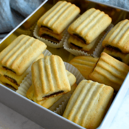 A tin box filled with rectangular date-filled cookies, each with a golden, ridged top and placed in white paper liners.