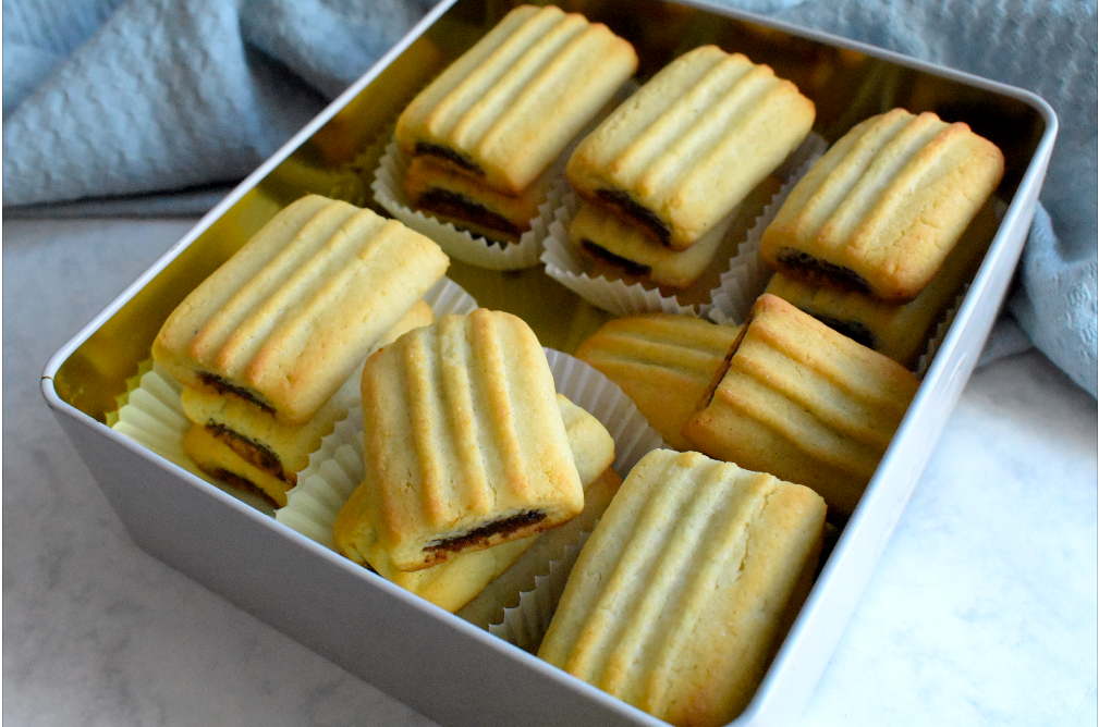 A tin box filled with rectangular date-filled cookies, each with a golden, ridged top and placed in white paper liners.
