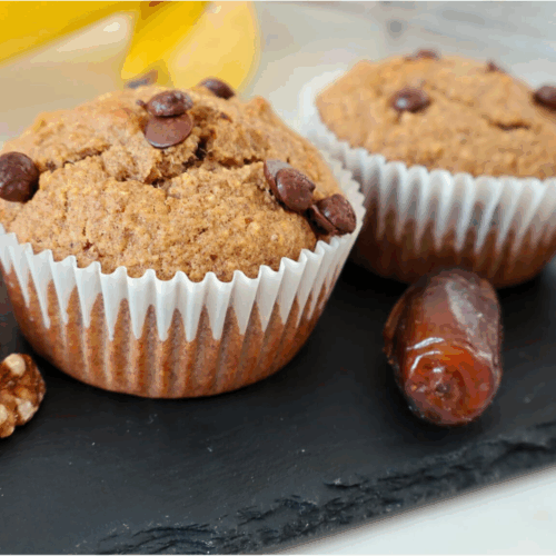 Three banana date muffins in a black plate, each in a clear cupcake liner, with whole dates and walnut halves placed beside them
