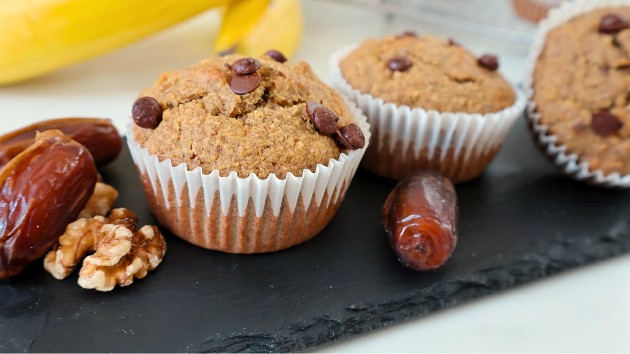 Three banana date muffins in a black plate, each in a clear cupcake liner, with whole dates and walnut halves placed beside them