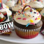Colorful rainbow cupcakes with layered inside, topped with whipped cream frosting, rainbow sprinkles, and rainbow candy pieces, displayed in cupcake liners on a white plate
