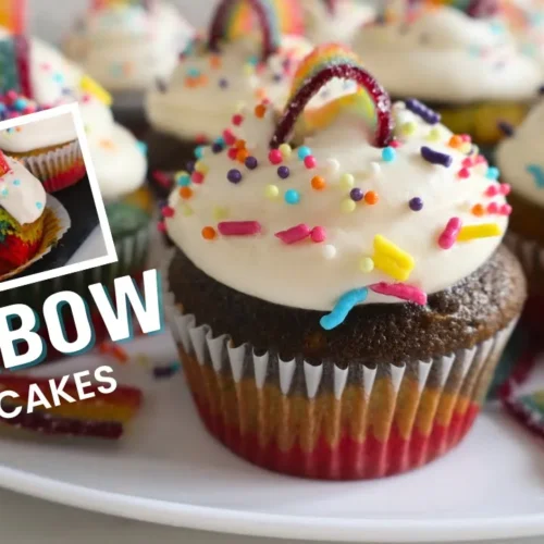 Colorful rainbow cupcakes with layered inside, topped with whipped cream frosting, rainbow sprinkles, and rainbow candy pieces, displayed in cupcake liners on a white plate