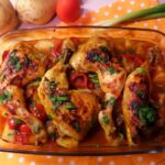 Baked chicken thigh casserole served with roasted potatoes, onion and sweet peppers, garnished with parsley, alongside a portion of steamed white rice topped with green onions. The dish is presented on a white plate, with the casserole dish full of chicken and vegetables in the background