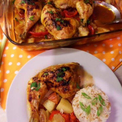Baked chicken thigh casserole served with roasted potatoes, onion and sweet peppers, garnished with parsley, alongside a portion of steamed white rice topped with green onions. The dish is presented on a white plate, with the casserole dish full of chicken and vegetables in the background