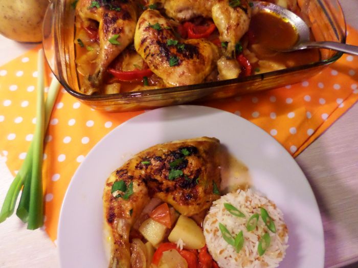 Baked chicken thigh casserole served with roasted potatoes, onion and sweet peppers, garnished with parsley, alongside a portion of steamed white rice topped with green onions. The dish is presented on a white plate, with the casserole dish full of chicken and vegetables in the background