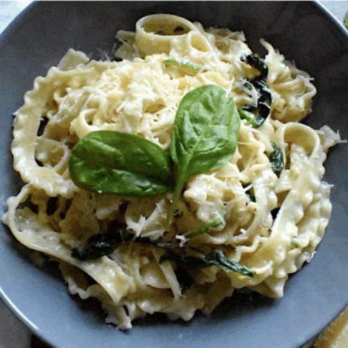 A bowl of 5-minute creamy spinach pasta served in a gray dish, topped with freshly grated Parmesan cheese and vibrant 2 spinach leaves. A fork and spoon rest beside the dish, with block of Parmesan cheese nearby for decoration
